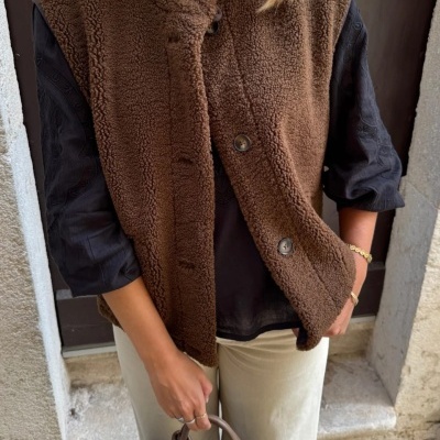 Colete castanho felpudo com botões vestido sobre camisa preta e calças claras, segurando bolsa castanha