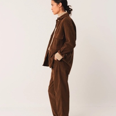 Mulher de perfil vestindo casaco e calças castanhas de veludo, camisola creme de malha e sapatos castanhos num fundo neutro.