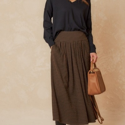 Mulher vestindo camisola preta, saia de padrão xadrez castanho e botas pretas em fundo bege.