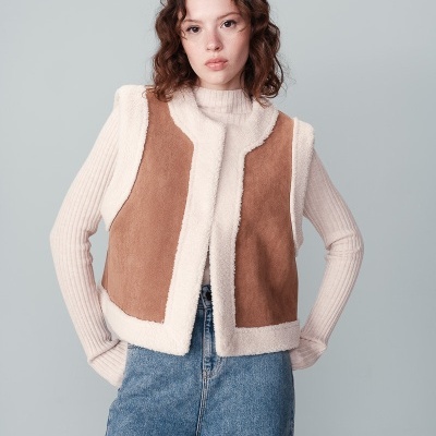 Colete castanho com forro branco por cima de camisola creme e calças de ganga.