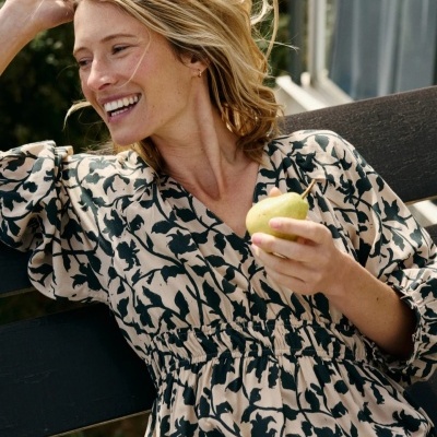 Mulher a sorrir sentada num banco preto a usar vestido floral bege e preto e a segurar uma pera verde