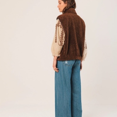 Mulher de costas com colete castanho felpudo, camisa creme bordada e calças largas azuis.