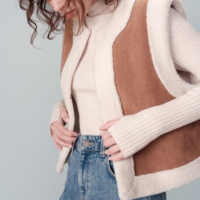 Mulher com colete castanho de camurça e pelo branco, camisola creme de gola alta e jeans azuis.