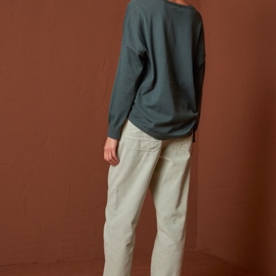 Pessoa de costas com camisola verde-escura, calças bege, meias transparentes e sapatos castanhos em fundo castanho