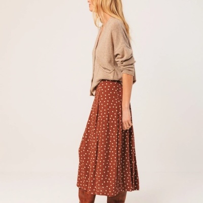 Mulher de lado com camisola bege, saia castanha com bolinhas e botas castanhas