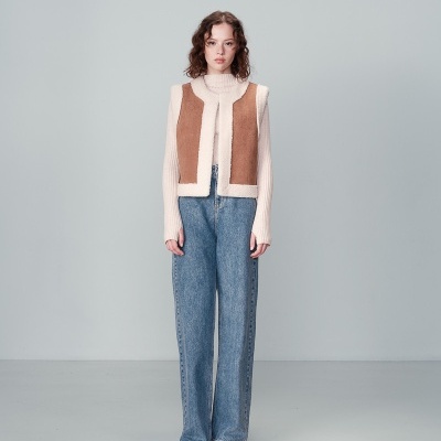 Modelo com colete castanho e branco, camisola de gola alta branca, calças de ganga azul e sapatos pretos num fundo cinza.