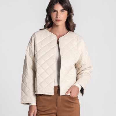 Mulher a usar casaco matelassé creme de padrão losangos, sobre camisola branca e calças castanhas.