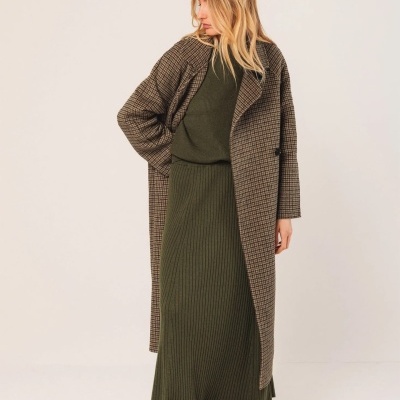 Mulher com casaco xadrez castanho e bege, camisola e saia verde escuro, botas castanhas, fundo creme