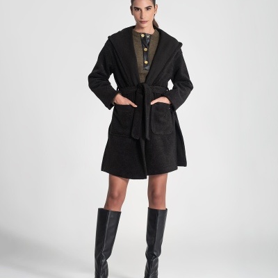 Modelo a usar casaco preto curto com capuz e cinto, vestido verde com botões dourados e botas pretas de couro altas.