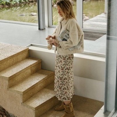 Mulher com casaco bege, saia floral e botas castanhas nas escadas interiores junto a janela grande