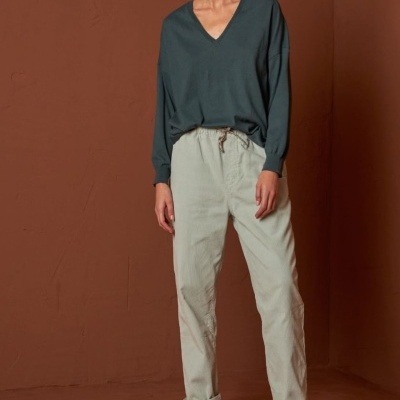 Mulher veste camisola verde, calças bege, meias bege e sapatos castanhos em fundo castanho.