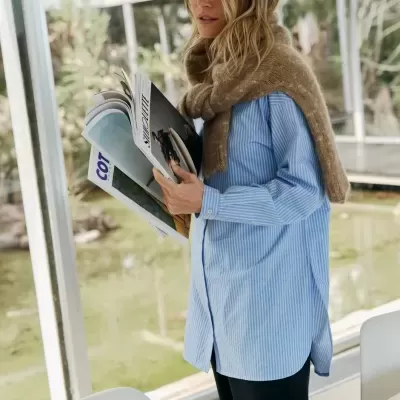 Mulher com camisa às riscas azuis e cachecol castanho segurando revistas.