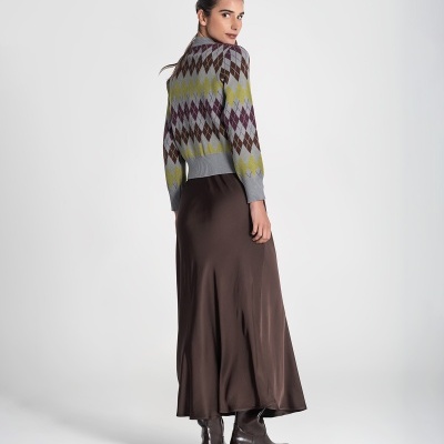 Mulher com camisola de malha geométrica, saia comprida castanha e botas castanhas em fundo branco.