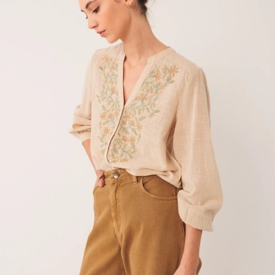 Blusa bege com bordado floral e calças castanhas em fundo branco
