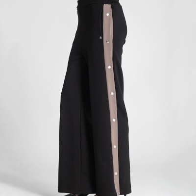 Calças pretas com risca lateral castanha e botões, cintura elástica, usadas com sapatos pretos de salto.