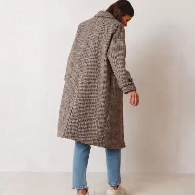 Casaco comprido xadrez bege e castanho usado por pessoa vista de costas, calça ganga azul clara e sapatilhas brancas.
