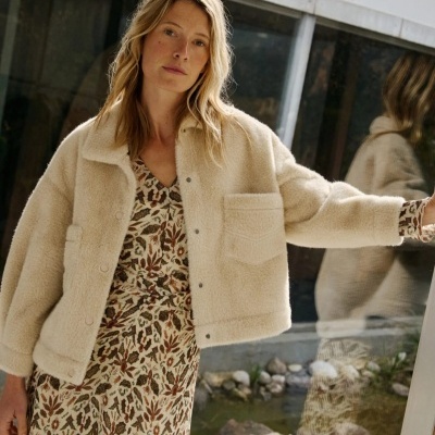 Mulher loira com casaco bege de pelo sintético e vestido floral em frente a vidro