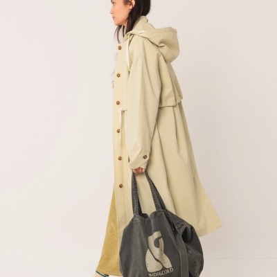 Mulher de perfil a usar casaco comprido bege com capuz, calças amarelas, ténis brancos e a segurar uma mochila cinzenta escura com logótipo e texto INDIGOOLD.