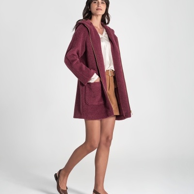 Mulher com casaco rosa escuro, blusa branca, saia castanha e sapatos baixos castanhos num estúdio branco.