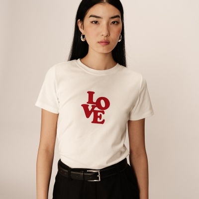 T-shirt branca com texto vermelho LOVE, usada por mulher com calças pretas, fundo neutro.