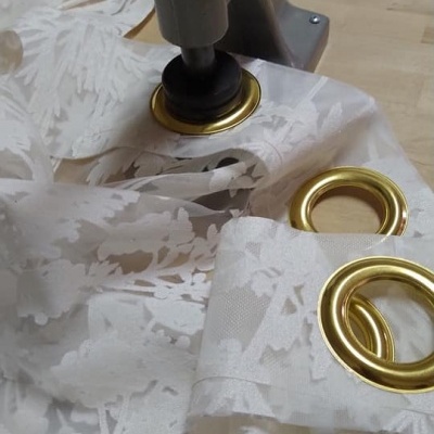 Tecido branco floral com ilhós dourados sendo aplicados por máquina