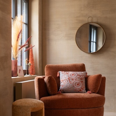 Cadeira castanho alaranjado com almofada decorativa e pufe numa sala com parede bege e espelho redondo