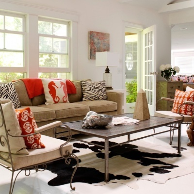 Sala de estar com sofá bege, almofadas zebras e laranja, cadeira estofada, mesa de metal e tapete branco e preto