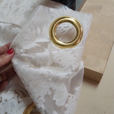 Tecido branco floral com ilhóses dourados e mão segurando sobre mesa com máquina.