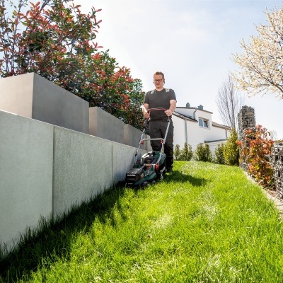 Homem a cortar relva com corta-relvas elétrico preto e verde num jardim entre muro branco e parede de pedra.