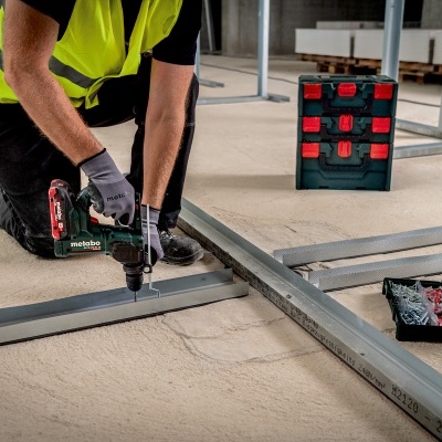 Pessoa com colete amarelo a usar furadeira eléctrica Metabo em estrutura metálica num chão cinzento.