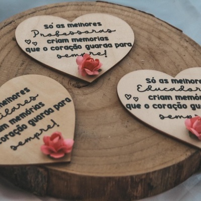 Três corações decorativos em madeira clara com texto em português e pequenas rosas cor-de-rosa, sobre uma base de tronco de madeira.