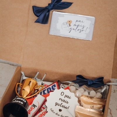 Caixa de presente com chocolates Kinder, troféu dourado, placa branca com mensagem e cartão com mensagem numa embalagem castanha com laço azul.
