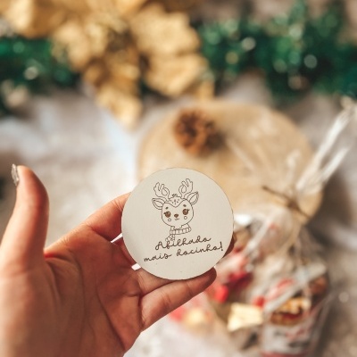 Etiqueta redonda branca com rena e texto Abilado mais docinho, mão segurando, fundo com decoração festiva e saco de doces desfocados