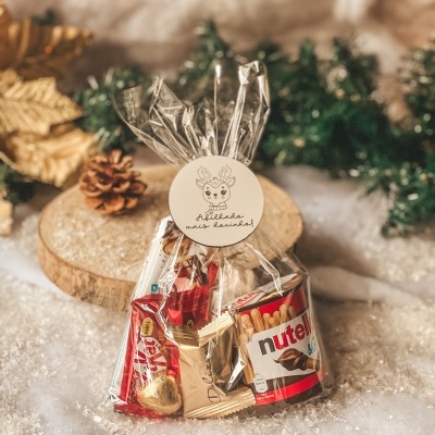 Embalagem transparente com doces variados e etiqueta com desenho de rena, numa superfície decorada para Natal.