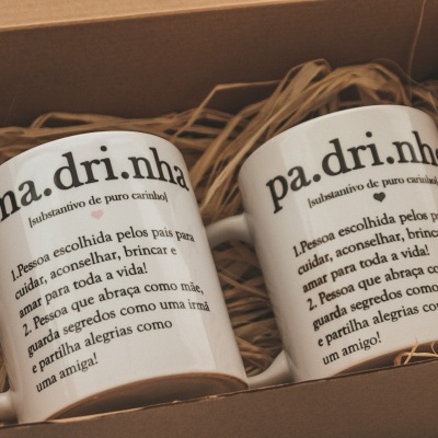Dois copos brancos com texto em português dentro de caixa de presente com palha artificial