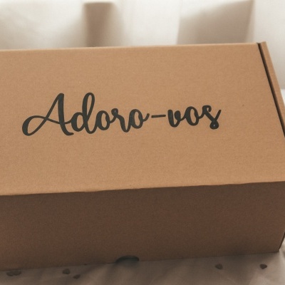 Caixa de cartão castanha com texto preto 'Adoro-vos' em superfície clara com flores