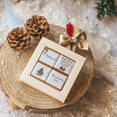 Caixa branca com quatro chocolates natalícios sobre base de madeira, com pinhões e laço dourado