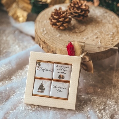 Conjunto de quatro chocolates embalados em caixa creme com fita dourada e etiquetas escritas em português, sobre tecido e madeira com pinhas