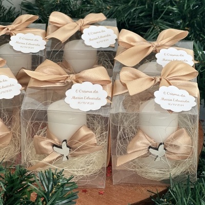 Caixas com velas brancas decoradas com laços bege e etiquetas personalizadas rodeadas por ramos verdes.
