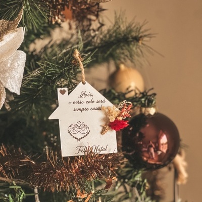 Enfeite de Natal em forma de casa branca pendurado numa árvore com bolas douradas e castanhas.