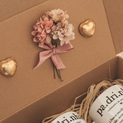 Caixa de cartão castanho com flores artificiais, dois frascos com texto em português e dois corações dourados