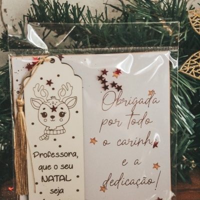 Marcapáginas e cartão de agradecimento natalício decorados com estrelas e rena em saco plástico sobre mesa com folhagem verde