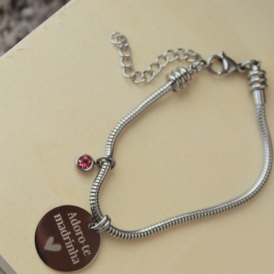 Pulseira prateada com pendente redondo castanho e pedra rosa sobre superfície de madeira clara