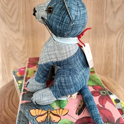 Urso de peluche azul e cinzento em tecido riscado com etiqueta e laço vermelho sentado sobre caixa floral colorida