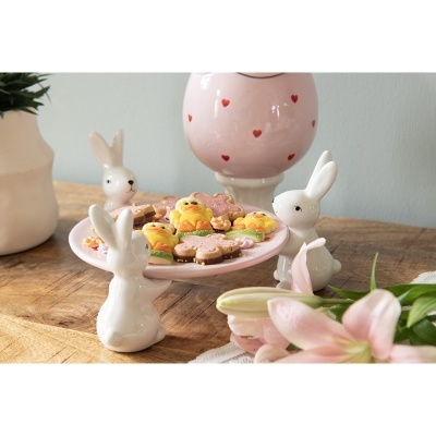 Prato decorativo branco com coelhos e biscoitos coloridos sobre mesa de madeira com jarra rosa e flores