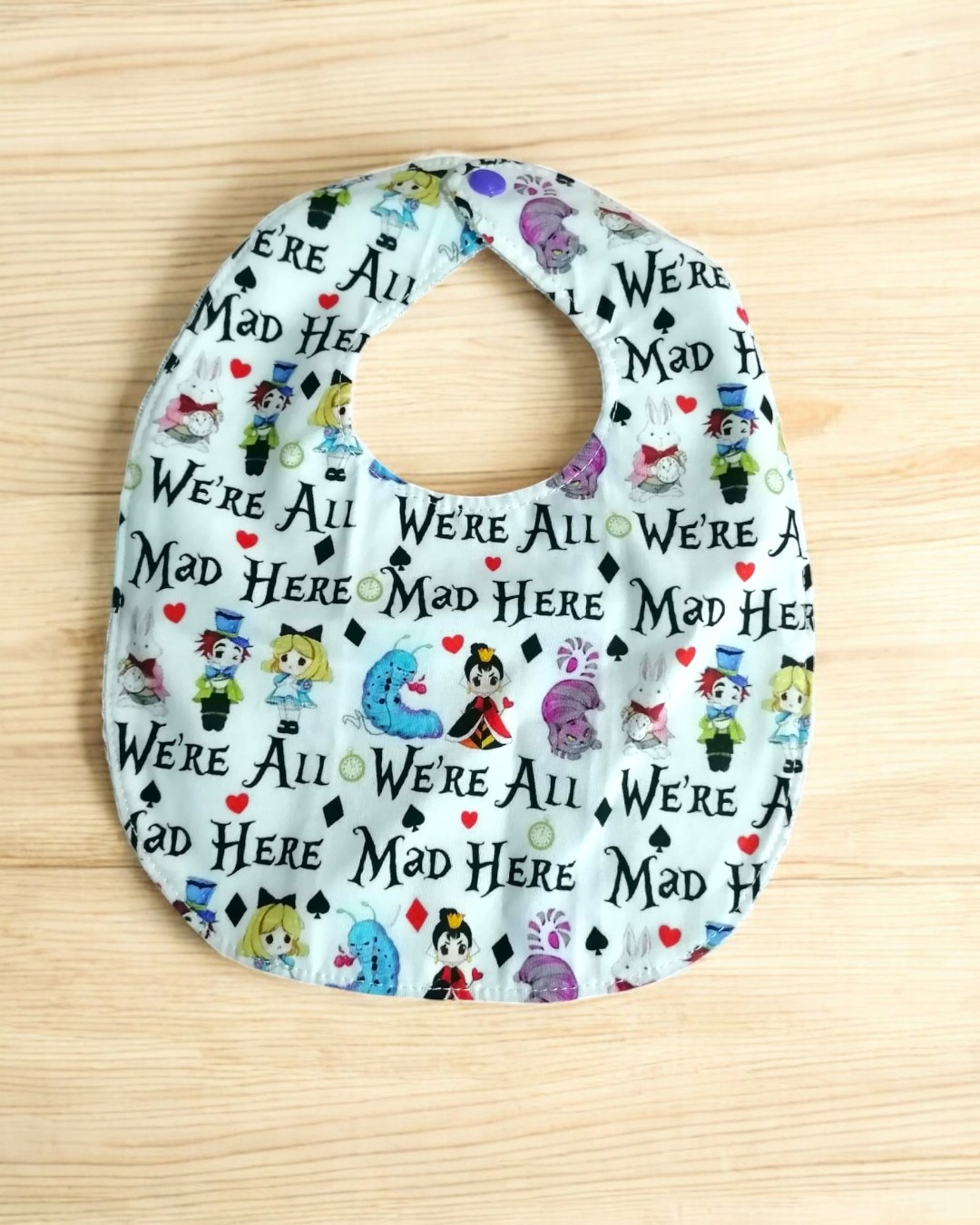 Babete branco com ilustrações de Alice no País das Maravilhas e texto 'WE'RE ALL MAD HERE' sobre fundo de madeira