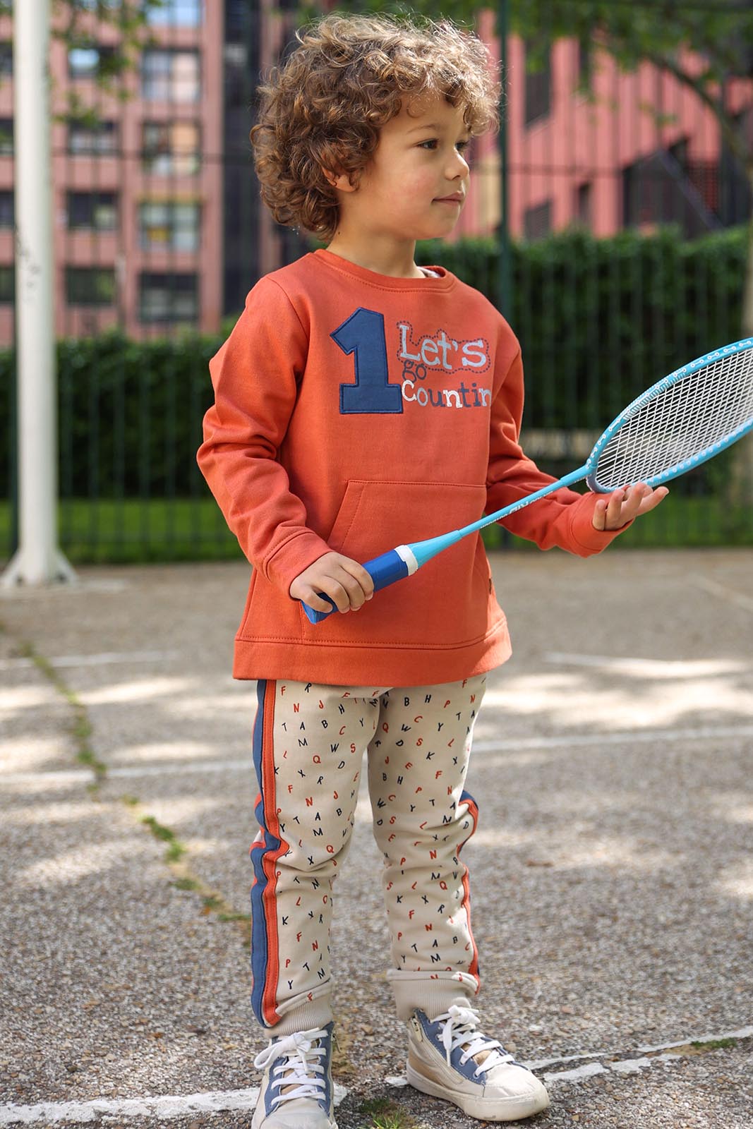 Criança com camisola laranja e calças bege com padrão de letras, a segurar raquete de ténis