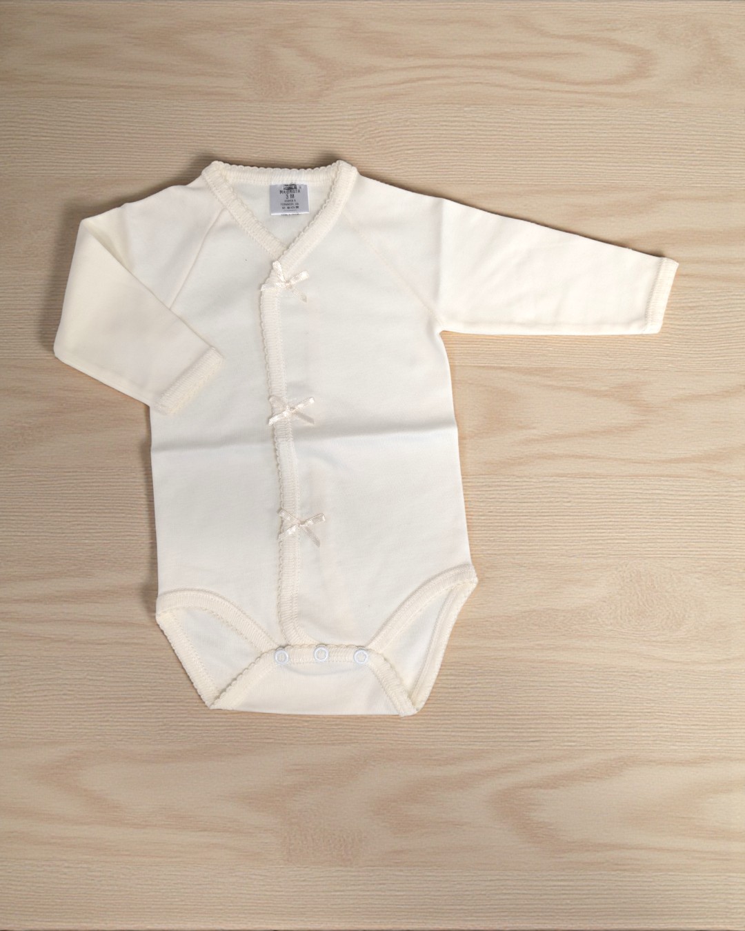 Body bebé creme de manga comprida com laços e molas sobre fundo madeira.