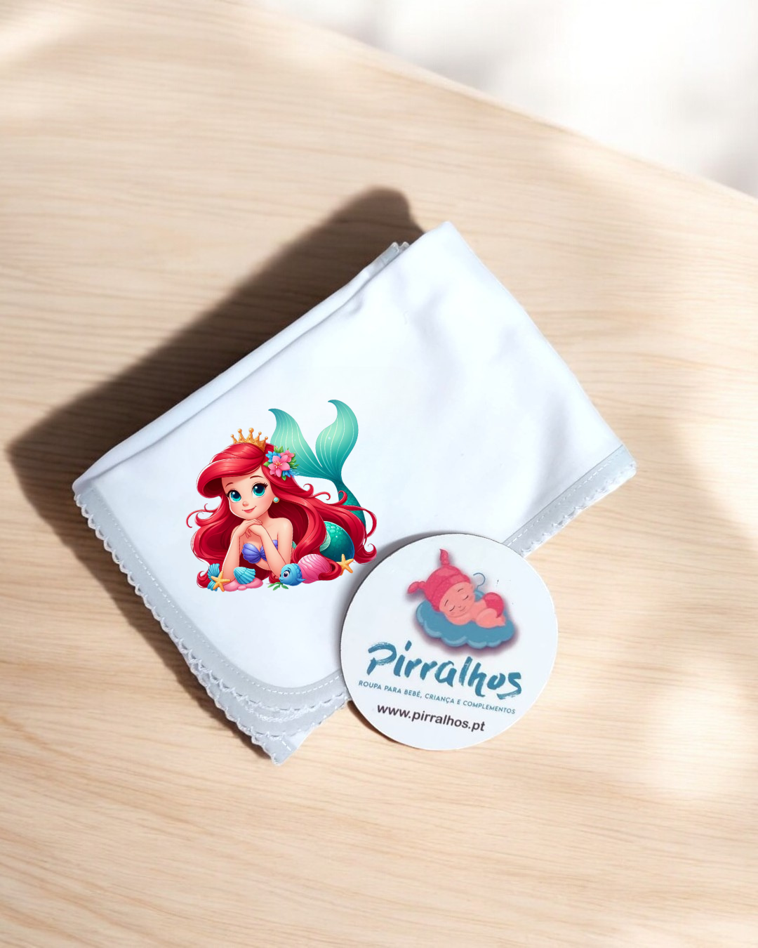 Lenço branco com estampa de sereia e etiqueta redonda de marca Pirralhos em superfície de madeira
