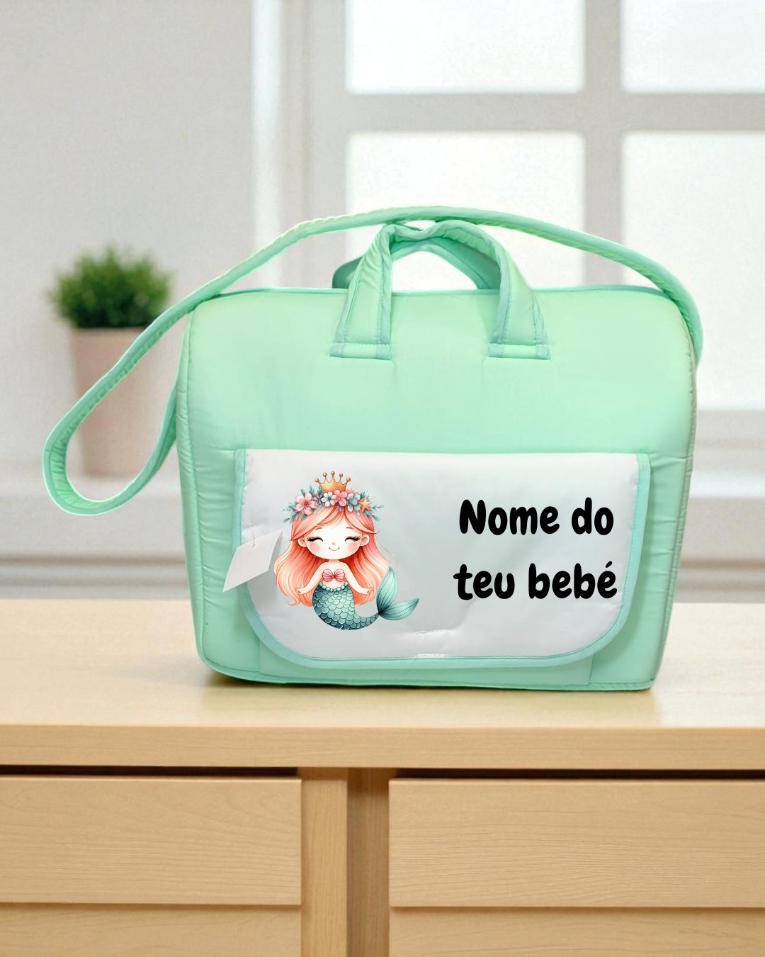 Bolsa para bebê verde água com bolso branco e desenho de sereia, texto 'Nome do teu bebé', sobre superfície de madeira